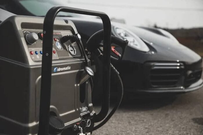 Krupni plan Idromatic Astra Vortex - Industrijski Parocistac (220°C, 3 operatera, dizel, 48kW) sa komandama i crijevom; elegantan crni auto u pozadini.