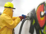 Noseći žuto zaštitno odijelo i kacigu, neko koristi Mosmatic FL-ABB 200 povrsinski cistac (200 mm, 120°C, 350 bara) za čišćenje šarenih grafita s bijelog zida.