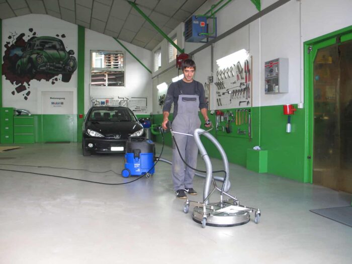 U dobro osvijetljenoj garaži, neko u sivom kombinezonu koristi Mosmatic FL-AHY 520 Površinski čistač. U blizini, mural zelenog automobila ukrašava zid pored parkiranog crnog automobila.