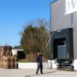 Sa teleskopskim stapom za čišćenje pod visokim pritiskom (275 bara, 100°C, 7,8m), osoba čisti industrijsku zgradu u blizini naslaganih paleta i utovarnog pristaništa.