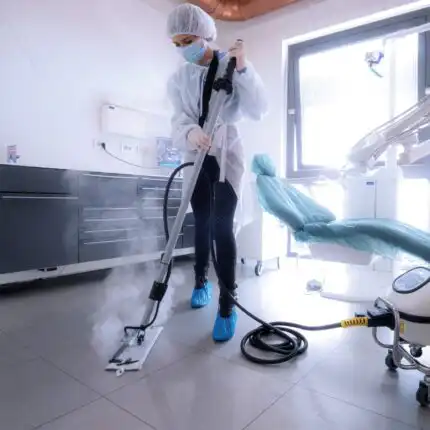U dobro osvijetljenoj prostoriji, vjerovatno stomatološkoj klinici, osoba u zaštitnoj opremi koristi parni čistač na podu; vidljiva je zubarska stolica.