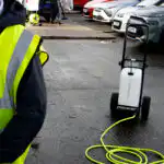 Na parkingu, osoba u dobro vidljivom prsluku stoji levo dok druga koristi Streamflo 25L sistem za pranje automobila, sa žutim crevom spremnim u blizini.