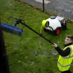 Osoba u neonskom zaštitnom prsluku čisti prozore pomoću šipke na izvlačenje i četke. U blizini, Streamflo 120L uređaj za čišćenje prozora sa rezervoarom za vodu, DI filterom 6,9L, litijumskom baterijom, pumpom od 6,9 bara i crevom od 50m stoji na travi pored trotoara i ograde na otvorenom.