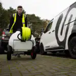 Osoba u dobro vidljivom prsluku gura kolica sa Streamflo 120L za čišćenje stakla, prolazeći pored parkiranih kombija, sa drvećem iza.