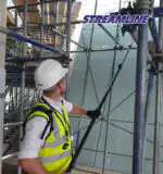 Radnik u belom šlemu i žutom prsluku vešto koristi alat na skelama, sa logom STREAMLINE na elegantnoj staklenoj fasadi iznad.