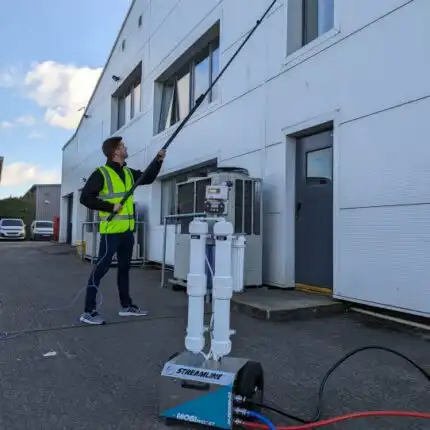 Osoba u neonskom prsluku čisti visoke prozore bele zgrade Mobi Midget 12V RODI sistemom, dok je mobilna mašina ispod.