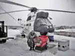 U zimskoj opremi, osoba stoji pored velikog helikoptera na snježnom asfaltu zagrijanom grijačem GE 36 Dizel Top, dok blagi snijeg pada.