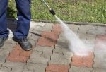 Osoba u tamnim cipelama i plavim pantalonama čiste šarene pločice Roto diznom, uz čistu površinu i travu sa žutim cvetovima.