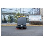 Mašina za čišćenje podova CHANCEE K100 stoji nepomično na asfaltu ispred staklene zgrade, sa drvećem i mutnim znakom iza nje.